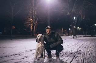 一个三十多岁的白人男子蹲在一只金发的拉布拉多猎犬旁边。他穿着一件绿色的冬季外套，手套，牛仔裤和棕色的鞋子。地上有雪，这是夜间。