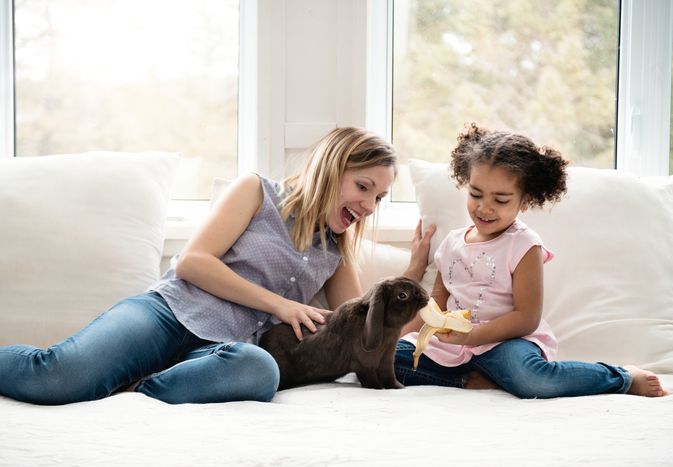 Rabbit eating a banana with a woman and little girl on a bed.